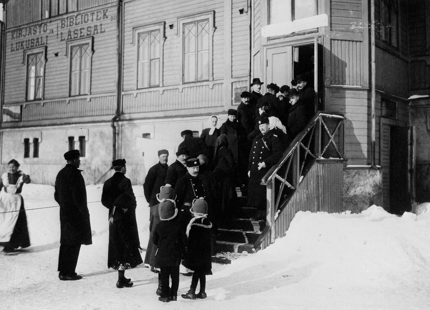 Valokuvassa äänestäjiä odottelemassa jonossa vuoroaan lumisessa talvisäässä kirjaston portailla Helsingissä. Kolme lasta etualalla katselemassa jonottajia. Kaksi vartijaa pitämässä järjestystä.