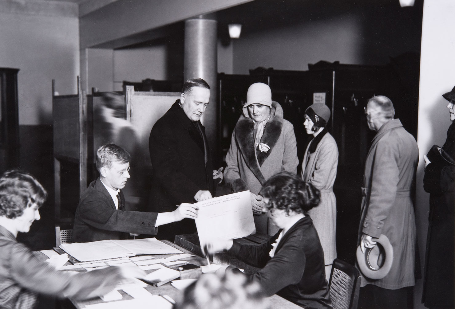 Valokuvassa Presidentti ja rouva Relander äänestyspaikalla presidentin valitsijamiesvaaleissa vuonna 1931. Kuvassa myös paikalla olevia vaalivirkailijoita sekä äänestäjiä jonottamassa vuoroaan.
