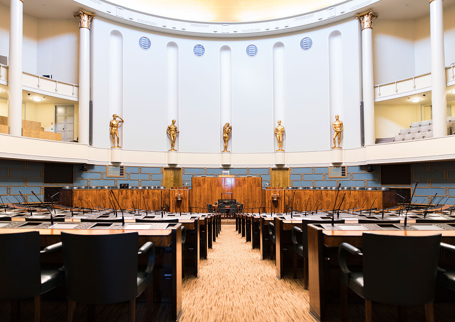 Valokuvassa yleiskuva nykyisestä eduskunnan istuntosalista tyhjillään. Keskellä istuntosalia koristavat patsaat, puheenjohtajien ja puhujan koroke. Kuvan sivustalta näkyvät myös yleisölehterit kahdessa kerroksessa.
